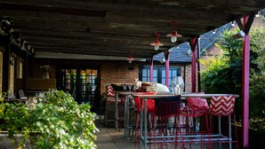 An outdoor patio area with metal tables and red chairs, a brick wall, and a covered section with hanging lights. - Huntsman of Brockenhurst (Brockenhurst)