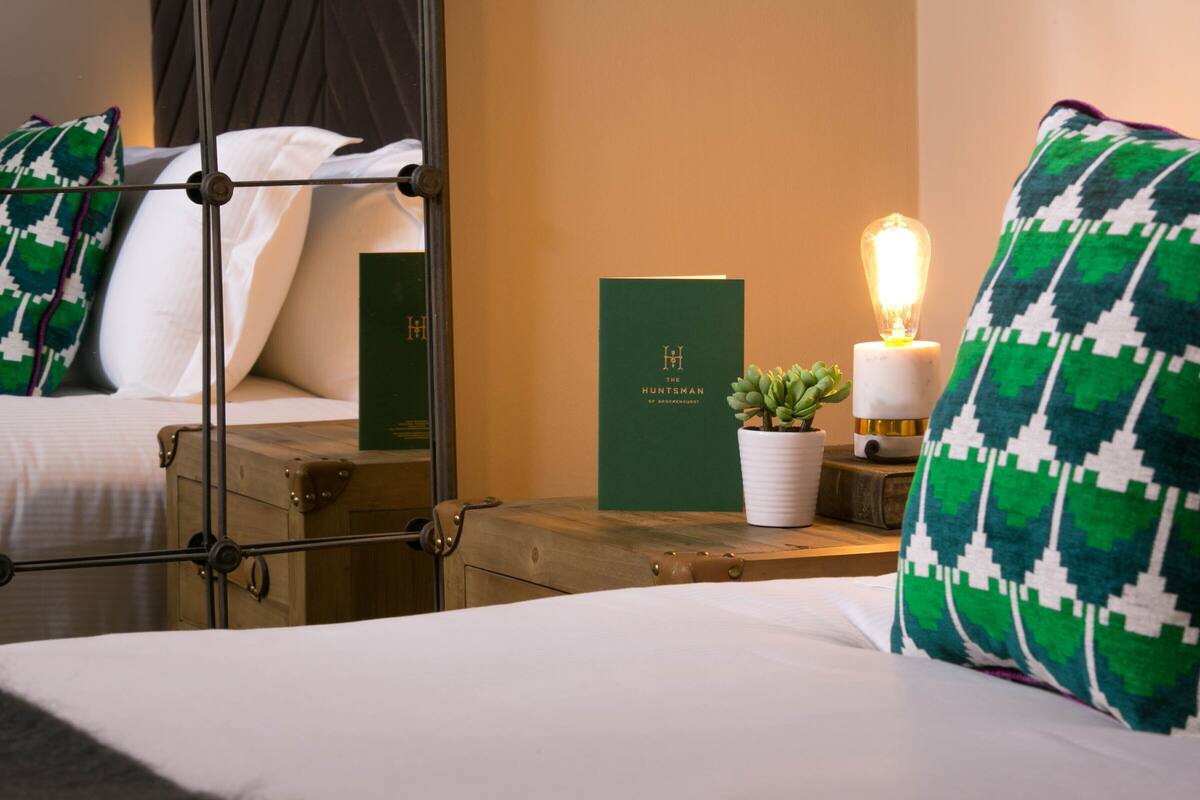 A hotel room with a bed, a bedside table with a book and a candle, a potted plant, and a suitcase.