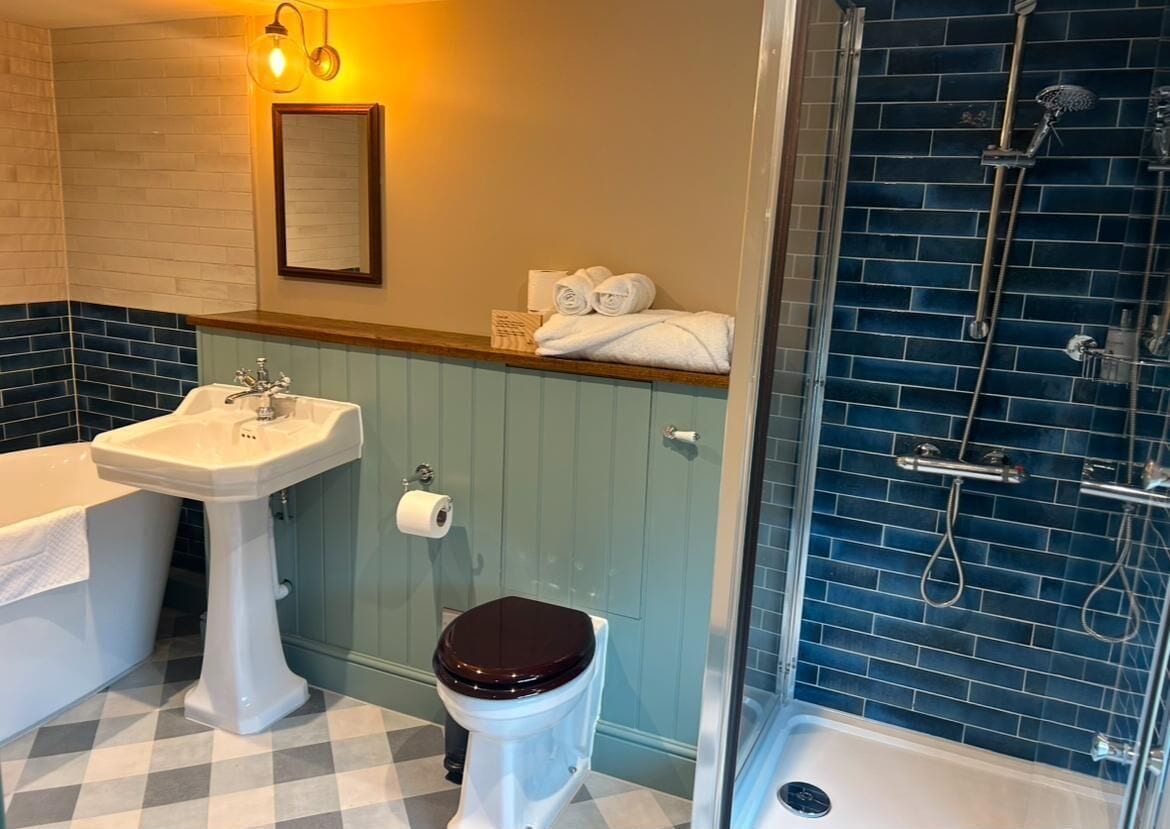 A bathroom with a white pedestal sink, a toilet with a wooden seat, a shower with a glass enclosure, and a blue-tiled shower wall.
