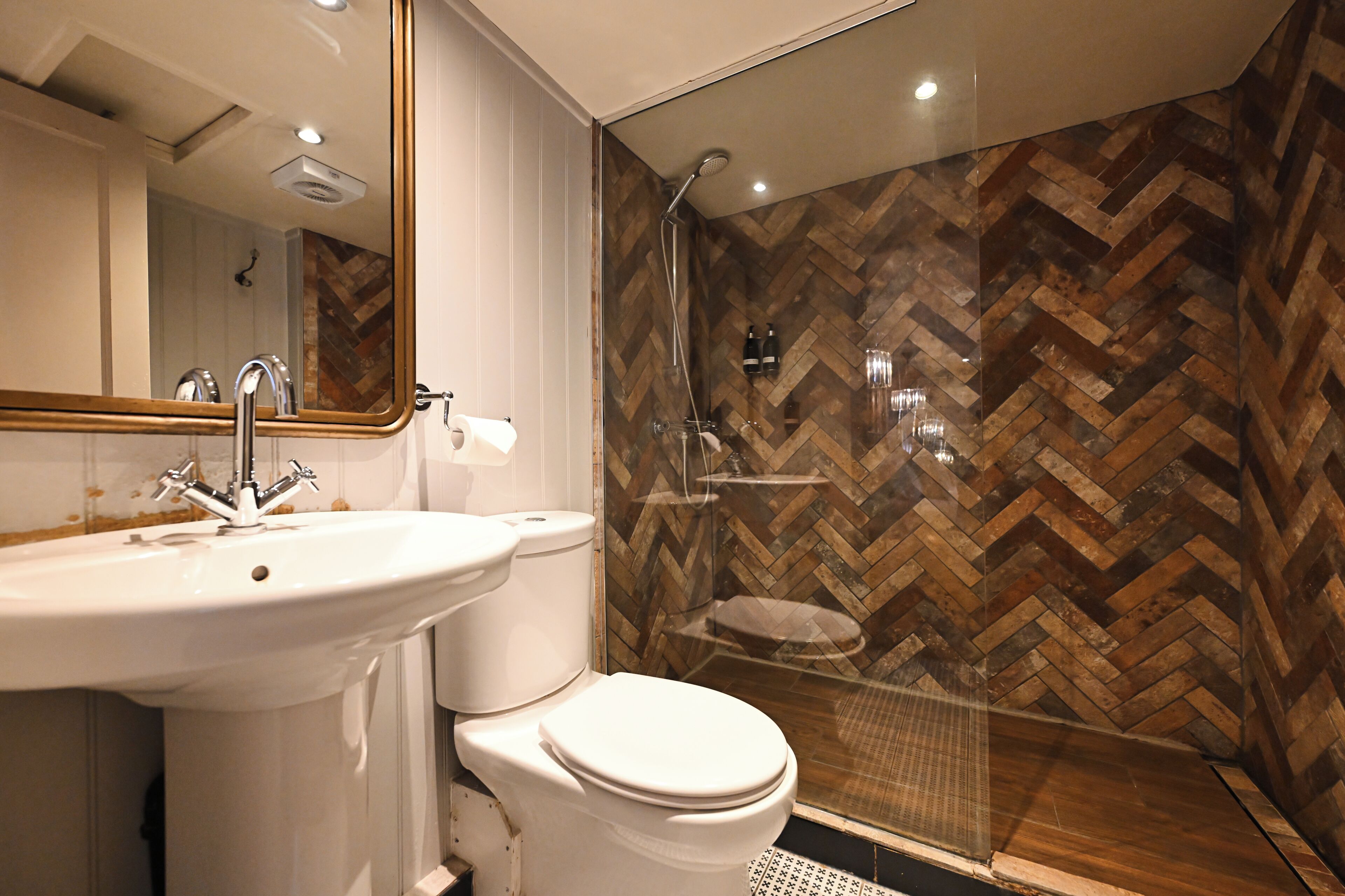 Un baño con pared de madera en patrón de espiga, un lavamanos blanco y una ducha con mampara de vidrio.