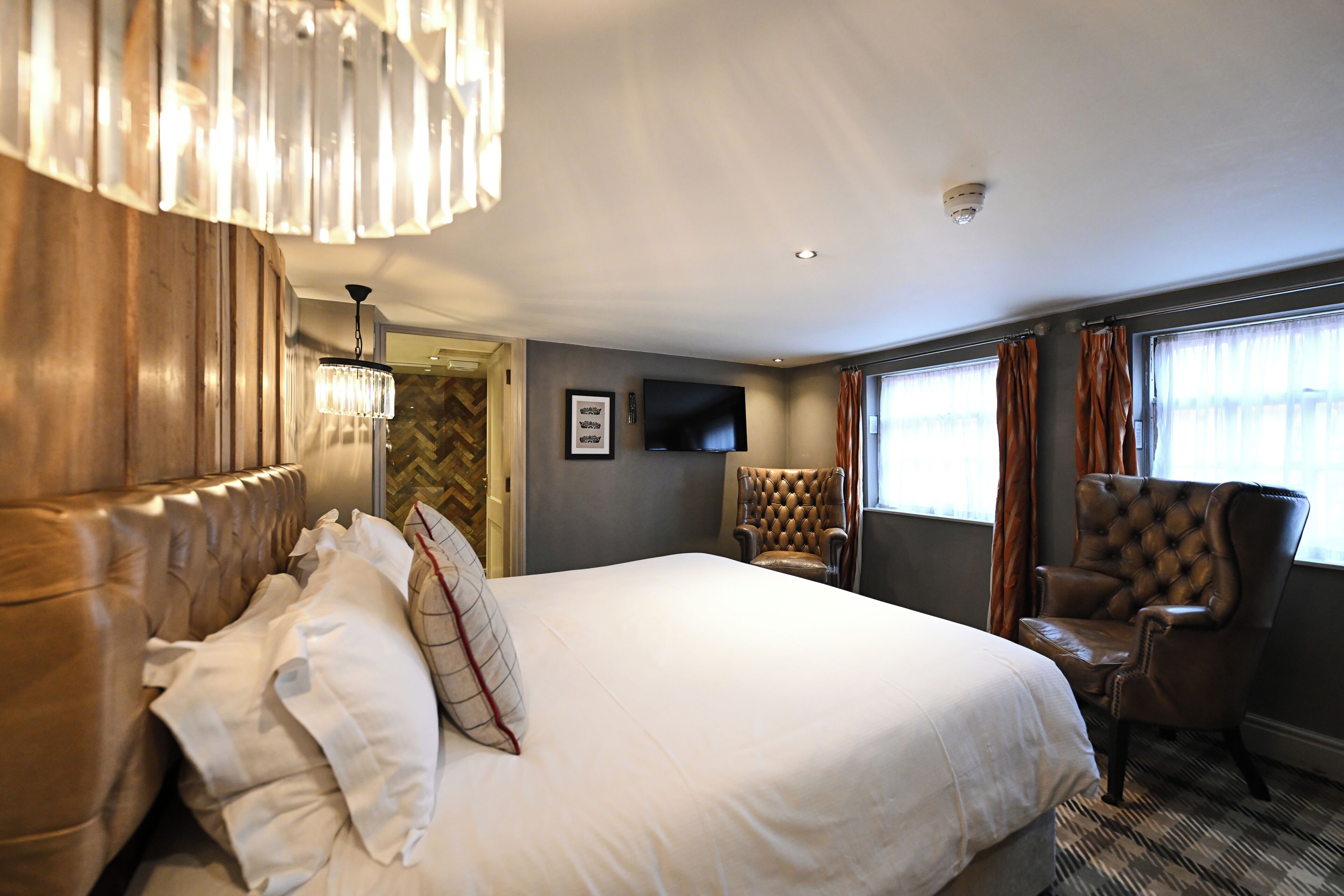A hotel room with a large bed, a tufted armchair, a television, and a chandelier.
