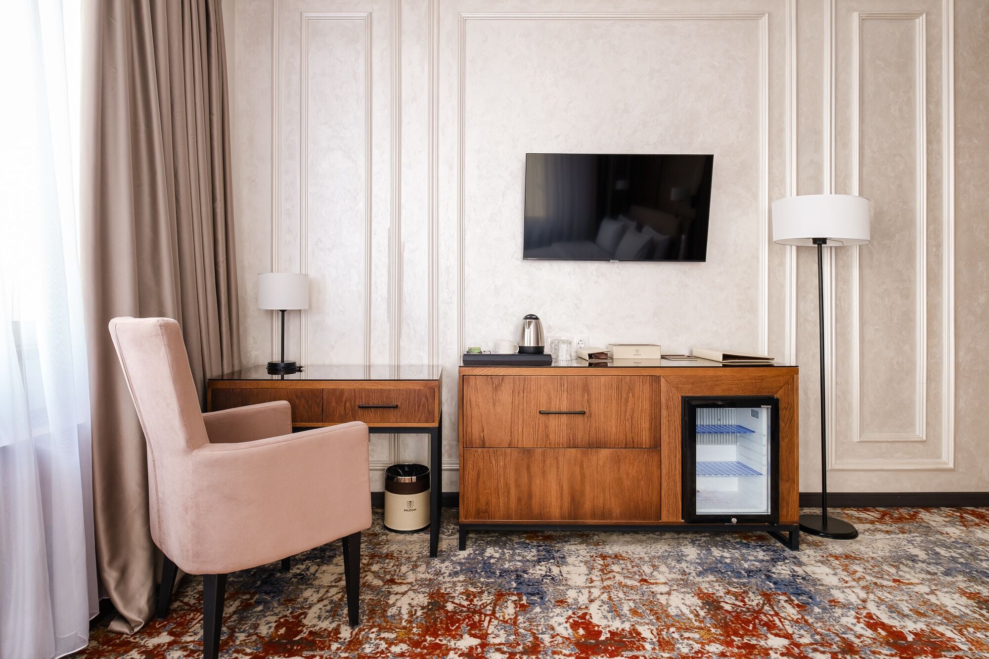 Quarto de hotel com escrivaninha de madeira, uma cadeira, televisão fixada na parede, abajur e uma lixeira.