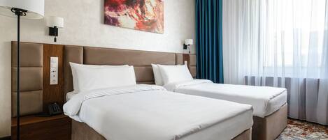 A hotel room with two beds, a headboard, bedside lamps, a power outlet, a telephone, a painting, and a window with curtains.