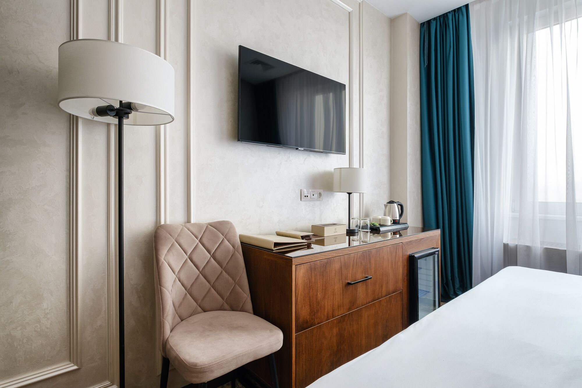 Habitación de hotel con cama, televisor montado en la pared, cómoda de madera, una silla y una lámpara.