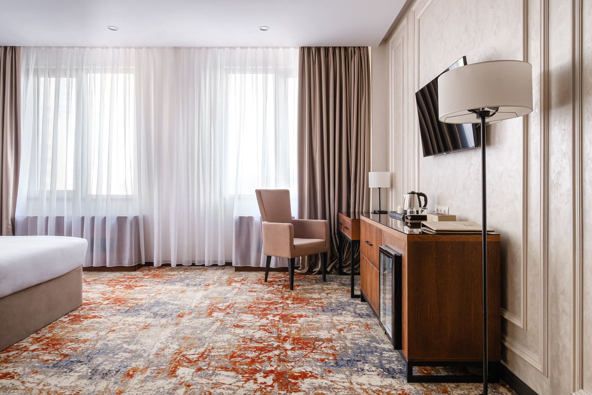 A hotel room with a bed, a desk with a lamp, a chair, and a TV mounted on the wall.