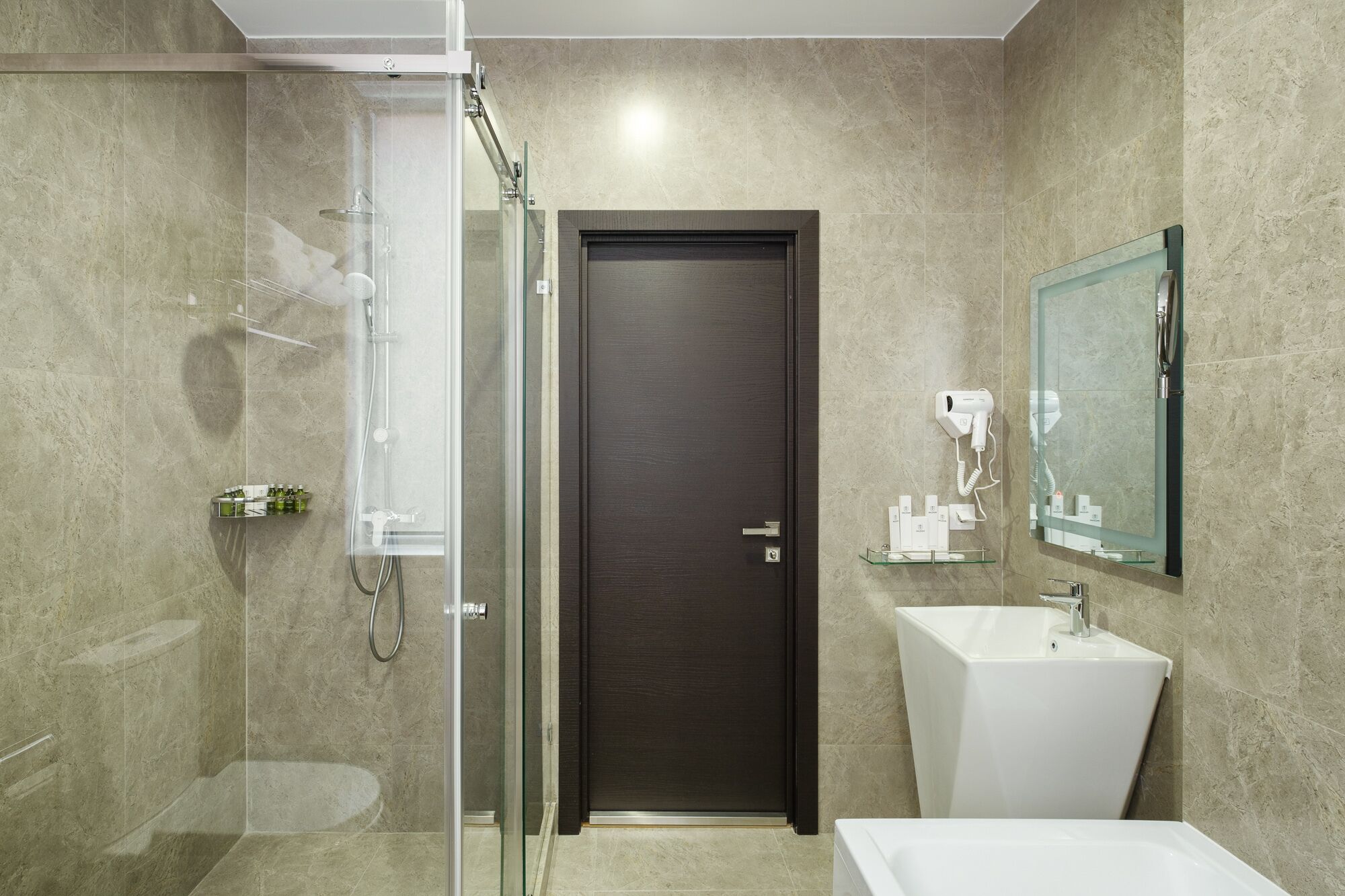 Un baño moderno con cabina de ducha de vidrio, una puerta de madera oscura, un lavamanos blanco y un espejo encima.