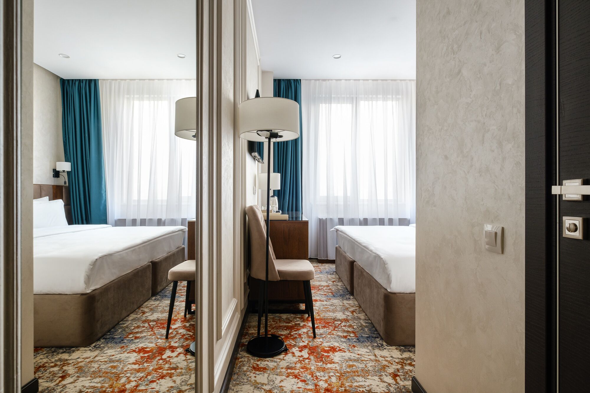 A hotel room with two beds, a desk, and a chair. There are two lamps on the desk and a large mirror on the wall.