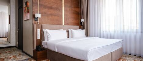 A hotel room with a large bed, a wooden accent wall, a telephone on the nightstand, and a view of the city through sheer curtains.