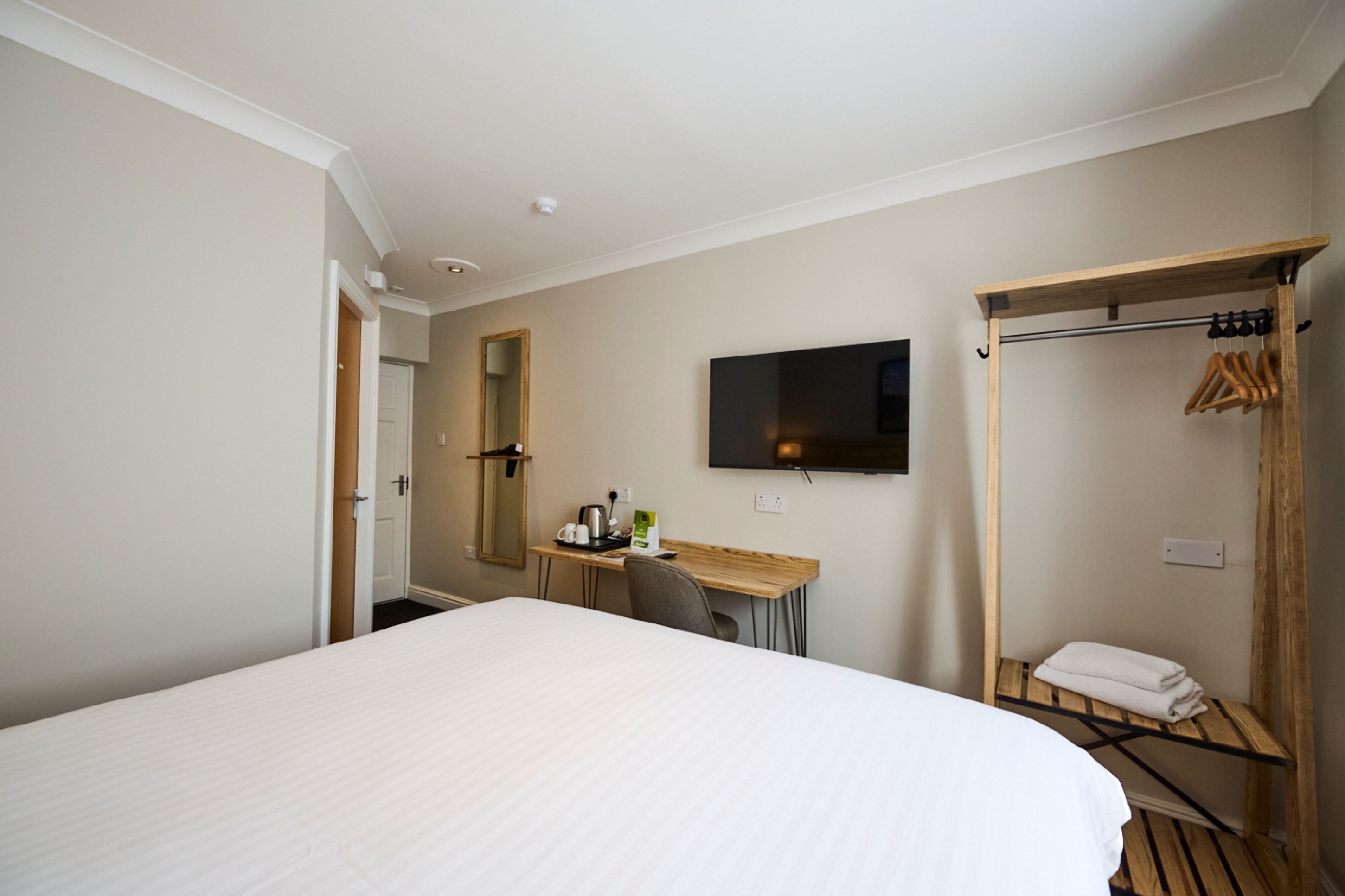 Habitación de hotel con una cama grande, un escritorio con silla, un televisor de pantalla plana en la pared y un perchero de madera con una toalla.