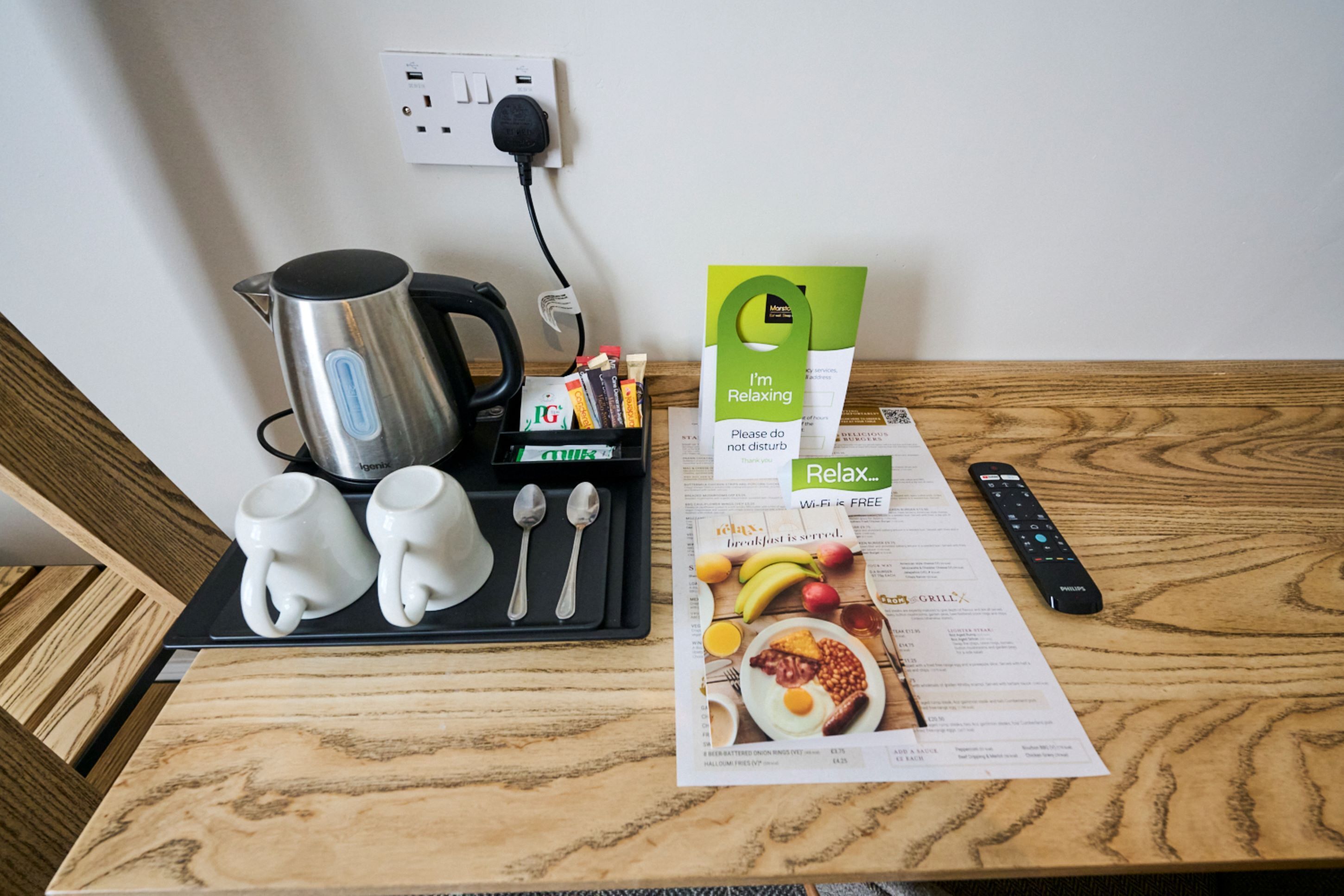 A wooden table with a kettle, mugs, spoons, a remote control, a menu, and a 'Relax' sign.