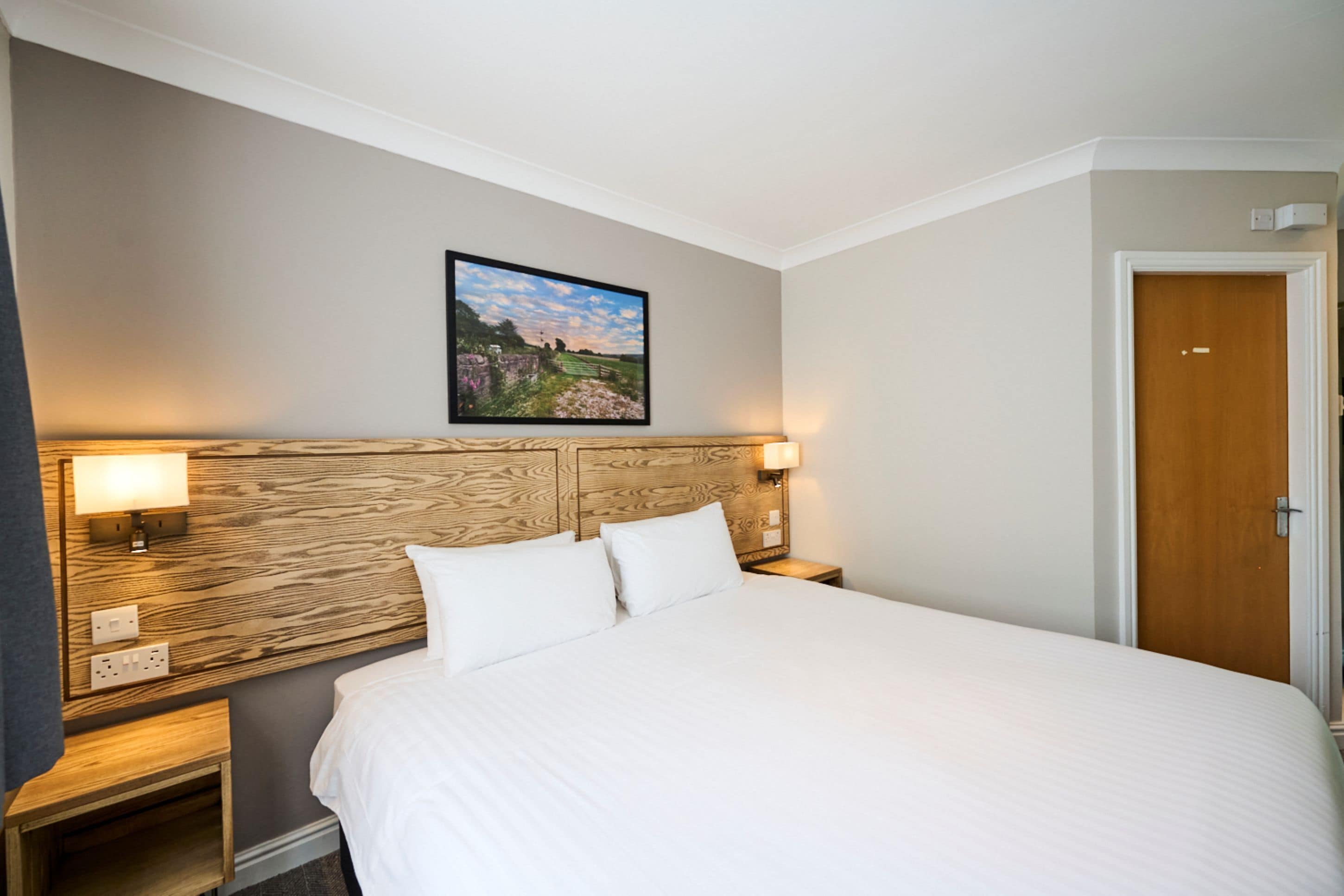 A hotel room with a large bed, wooden headboard, two bedside lamps, a painting on the wall, and a door to the right.