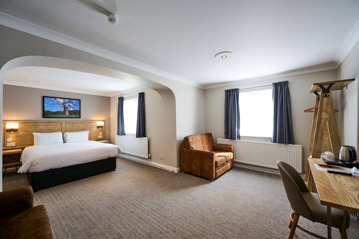 A hotel room with a bed, a seating area with a chair and table, a radiator, and a painting on the wall.