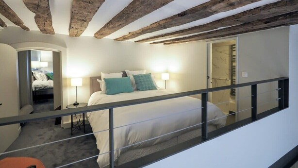 A bedroom with a bed, bedside lamps, a wooden ceiling, and a view of another room.