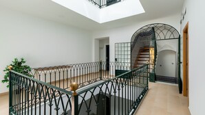 Un intérieur spacieux avec un plafond haut, un balcon doté d’une balustrade en verre, un escalier avec des rampes en métal décorées, et une vue sur un mur orné de motifs.