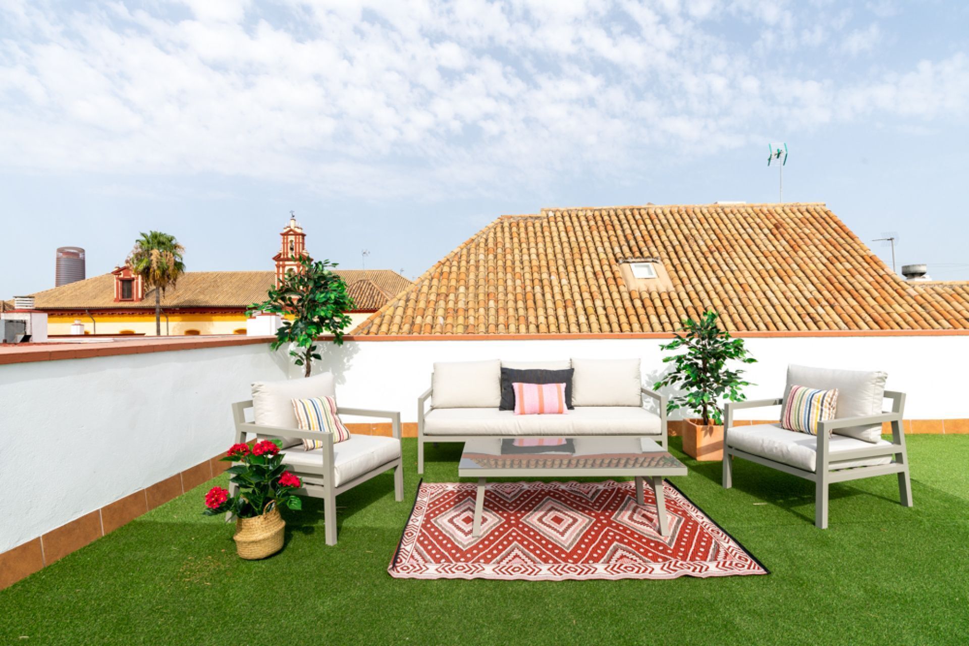 An outdoor seating area with a white sofa, two armchairs, a patterned rug, and potted plants.