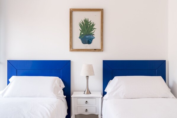 A hotel room with two beds, a nightstand, a lamp, and a framed botanical illustration on the wall.