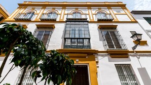 A building with a white facade and yellow trim, featuring multiple windows with metal bars, a central large window, and a door with a number plaque.