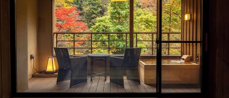 A room with a view of autumn trees through a large window, a small table with chairs, and a hot tub.