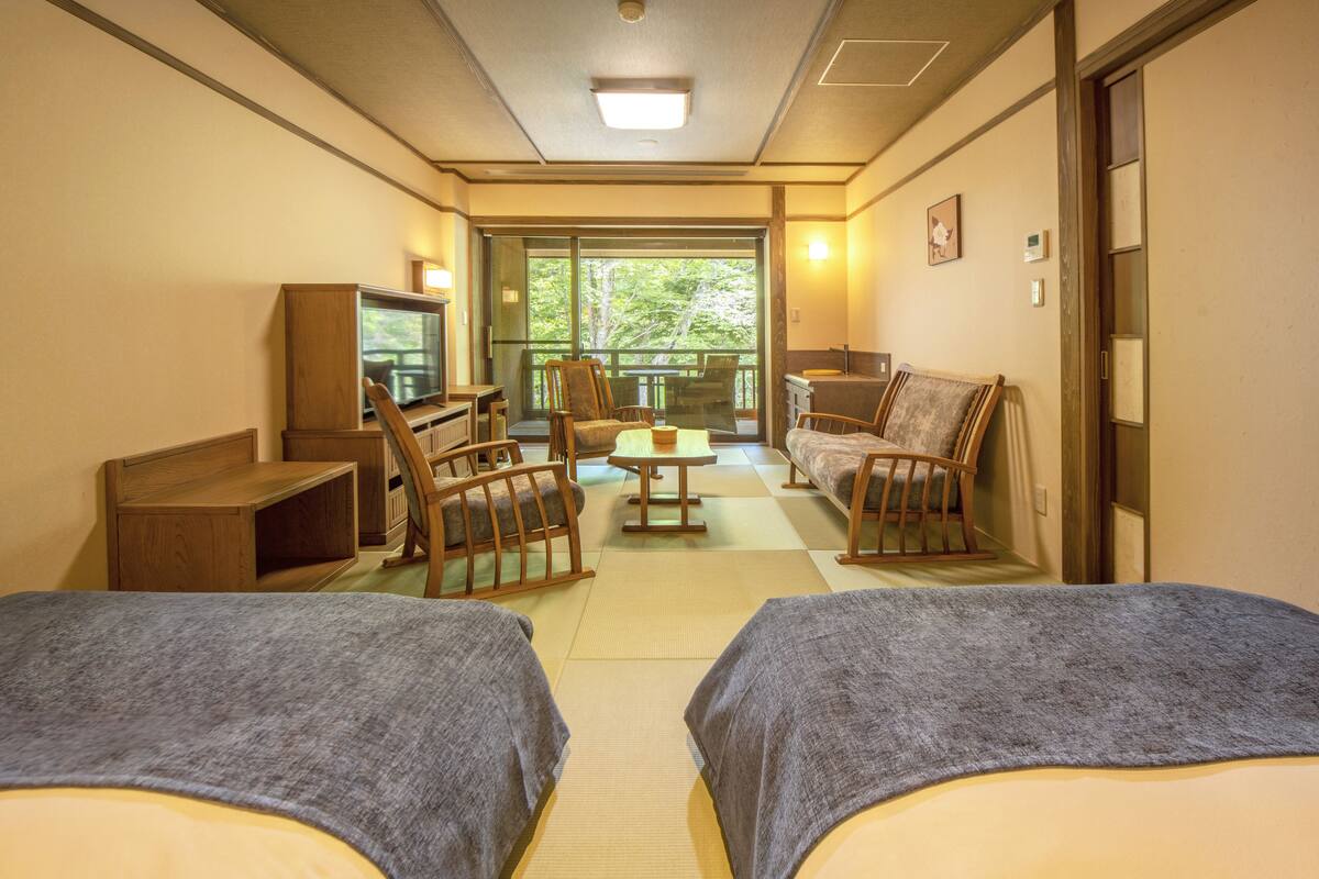 A hotel room with two beds, a TV, a sofa, and a balcony with a view of trees.
