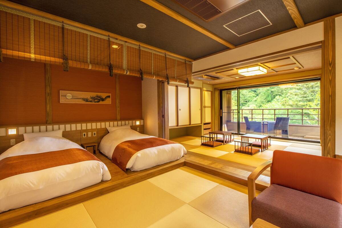 A traditional Japanese-style hotel room with tatami flooring, two beds, a wooden ceiling, and a view of greenery through large windows.