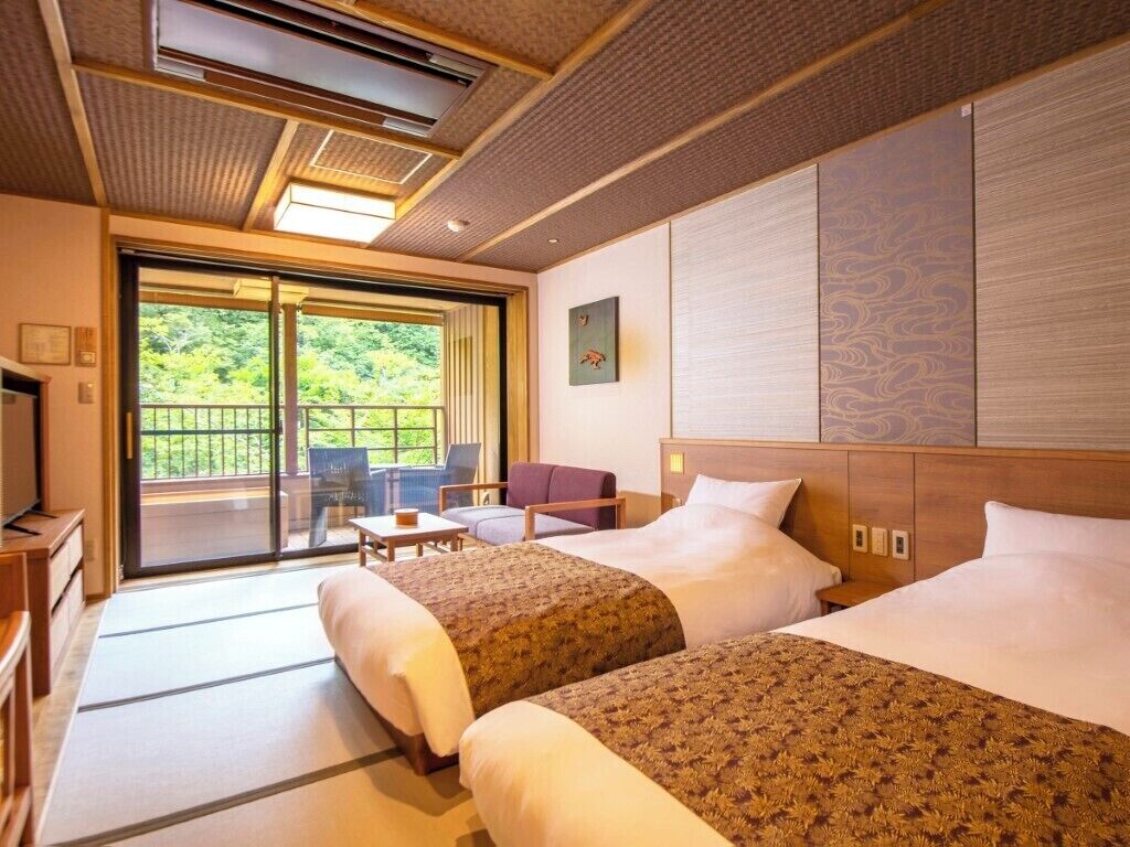 A hotel room with two beds, a sliding glass door to a balcony, a small table, and a chair.