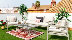 Terraza en la azotea con un sofá blanco, dos sillones, una mesa de centro de vidrio y un tapete estampado.