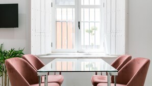 Una habitación con una mesa de vidrio y cuatro sillas rosas, una planta en maceta y una ventana con contraventanas blancas.