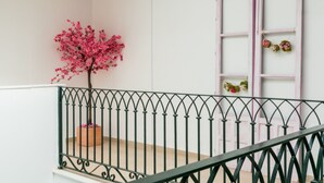 Un balcón con barandilla de hierro forjado, un árbol de flores rosas y una ventana con marco rosa adornada con un arreglo floral.