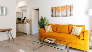 Ein modernes Wohnzimmer mit einem hellorangenen Sofa, einem Glastisch und einer Küche im Hintergrund.