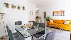 Ein modernes Wohnzimmer mit einem Esstisch aus Glas, grauen Stühlen, einem orangefarbenen Sofa und einer Küche im Hintergrund.