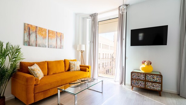 Ein Wohnzimmer mit einem senffarbenen Sofa, einem Glastisch, einem kleinen Holznachttisch mit einer gelben Vase und einem an der Wand befestigten Flachbildfernseher.