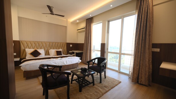 A modern hotel room with a large bed, a desk with a chair, a ceiling fan, and a view of the city through large windows. - Fairvacanze Inns & Suites Lucknow (Lucknow)