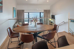 Un vaste espace dédié à la salle à manger, avec une table ronde en bois entourée de six chaises, un vase rempli de fleurs et une vue sur la mer grâce à de grandes fenêtres.