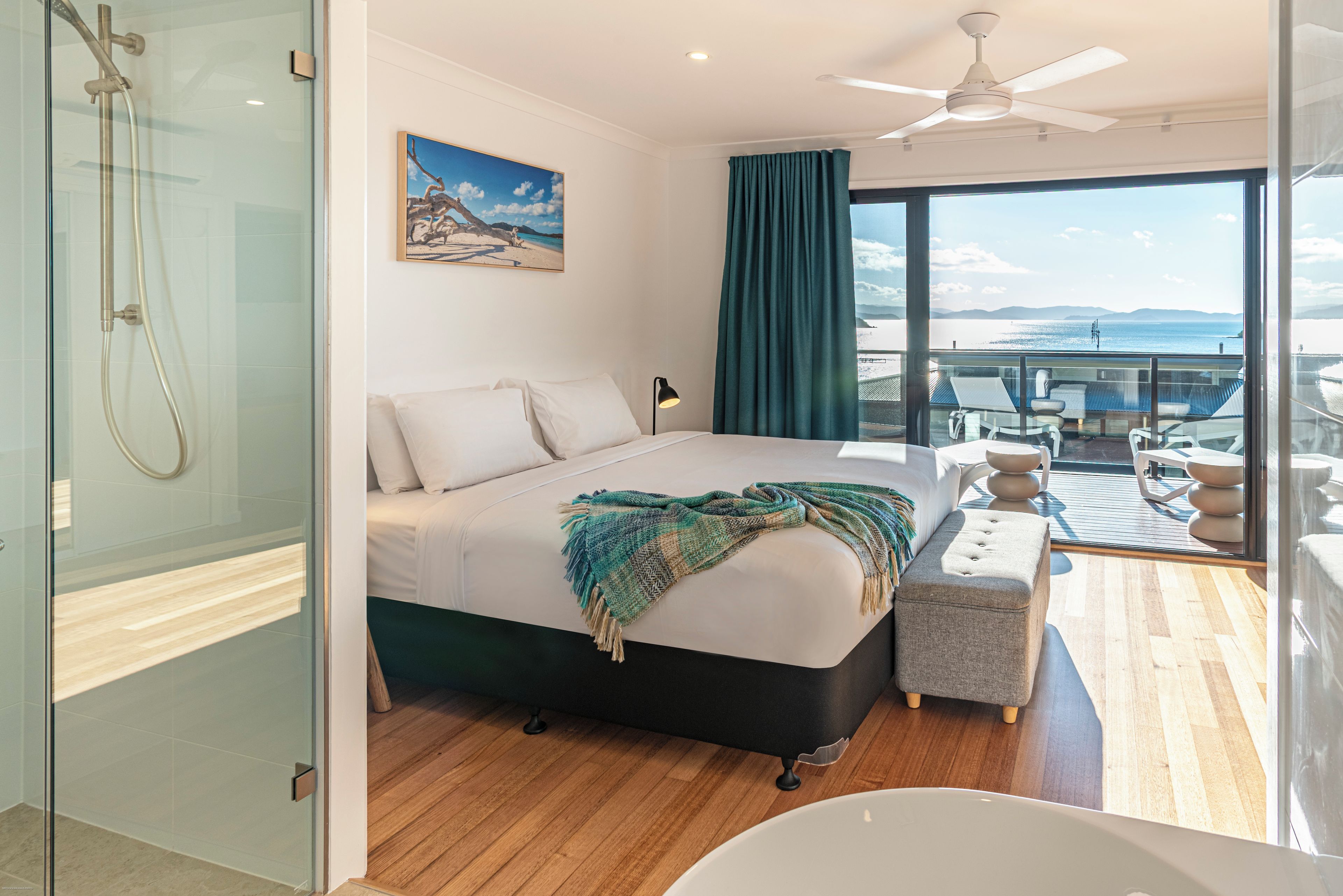 Un dormitorio moderno con una cama grande, vista al mar, un balcón con mobiliario exterior y una zona de ducha con paredes de cristal.