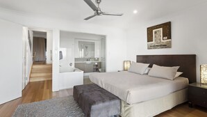 A modern bedroom with a large bed, a ceiling fan, a bedside table with a lamp, and a bathroom visible through an open door. - Shorelines 27 Hamilton Island - oceanviews, golf buggy and transfers (Hamilton Island)