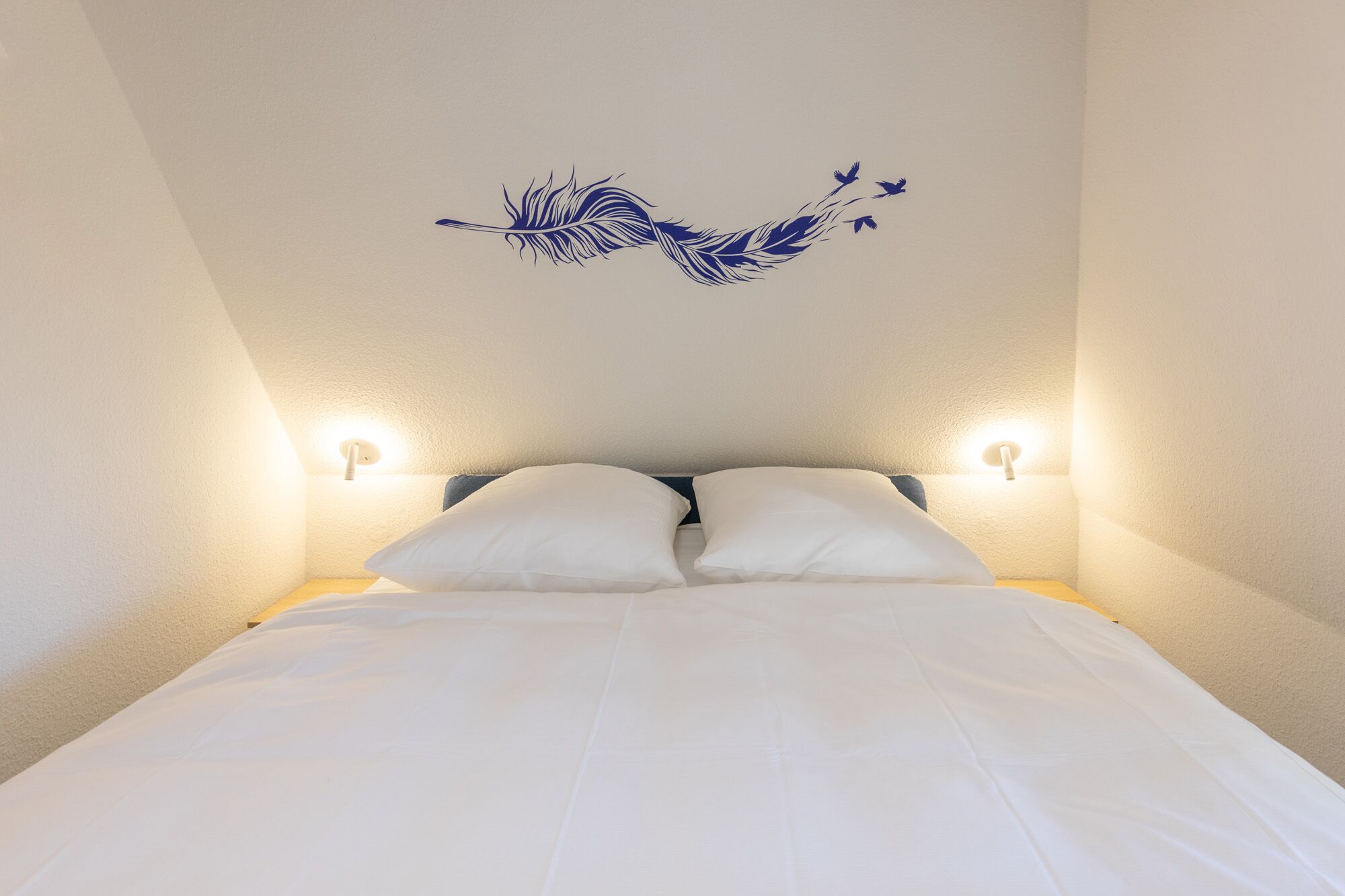 A bedroom with a bed, two bedside lamps, and a wall decal of a feather and birds.