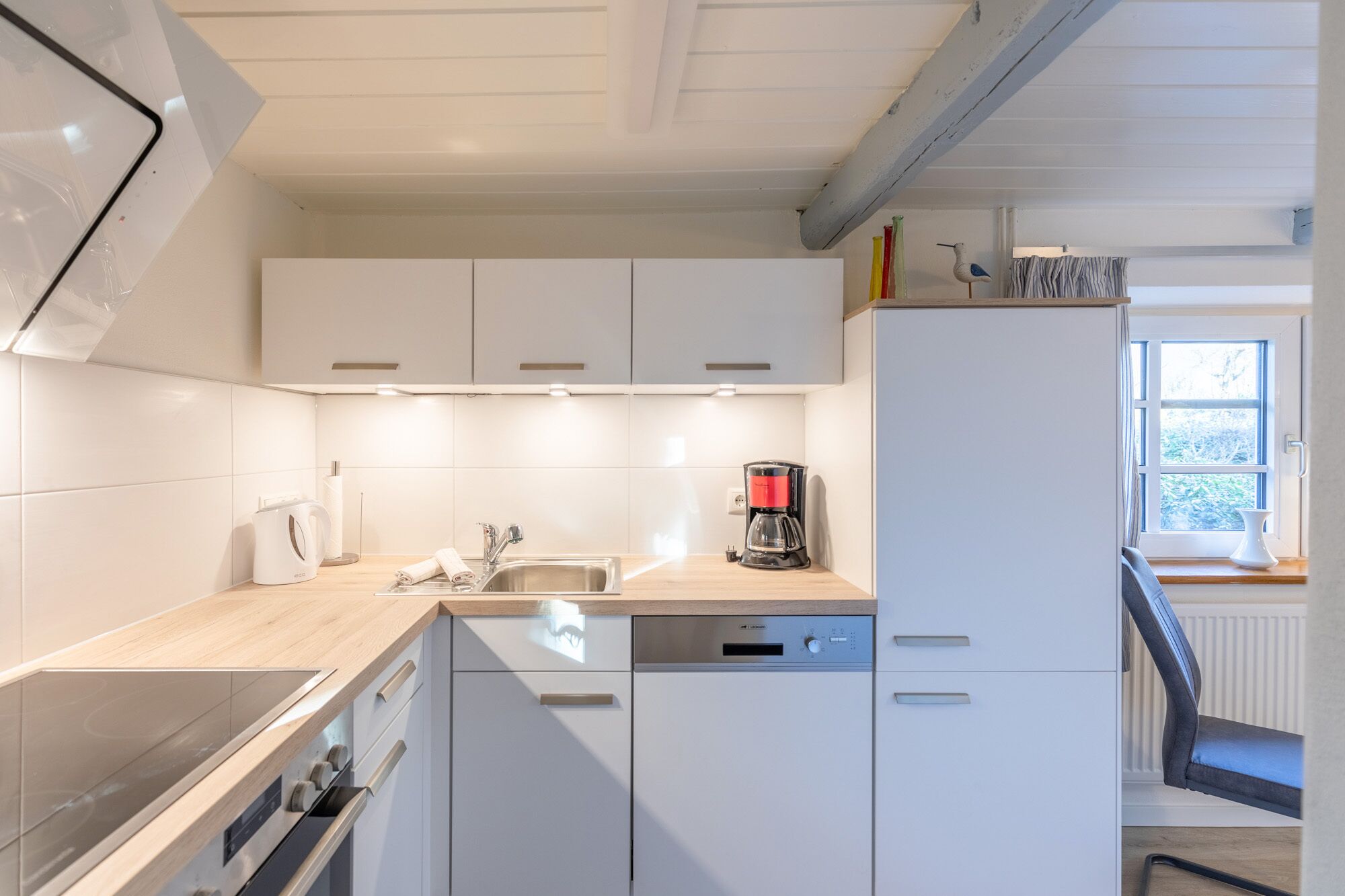 Une cuisine moderne avec des meubles blancs, un lave-vaisselle encastré, une machine à café et une fenêtre avec des rideaux.