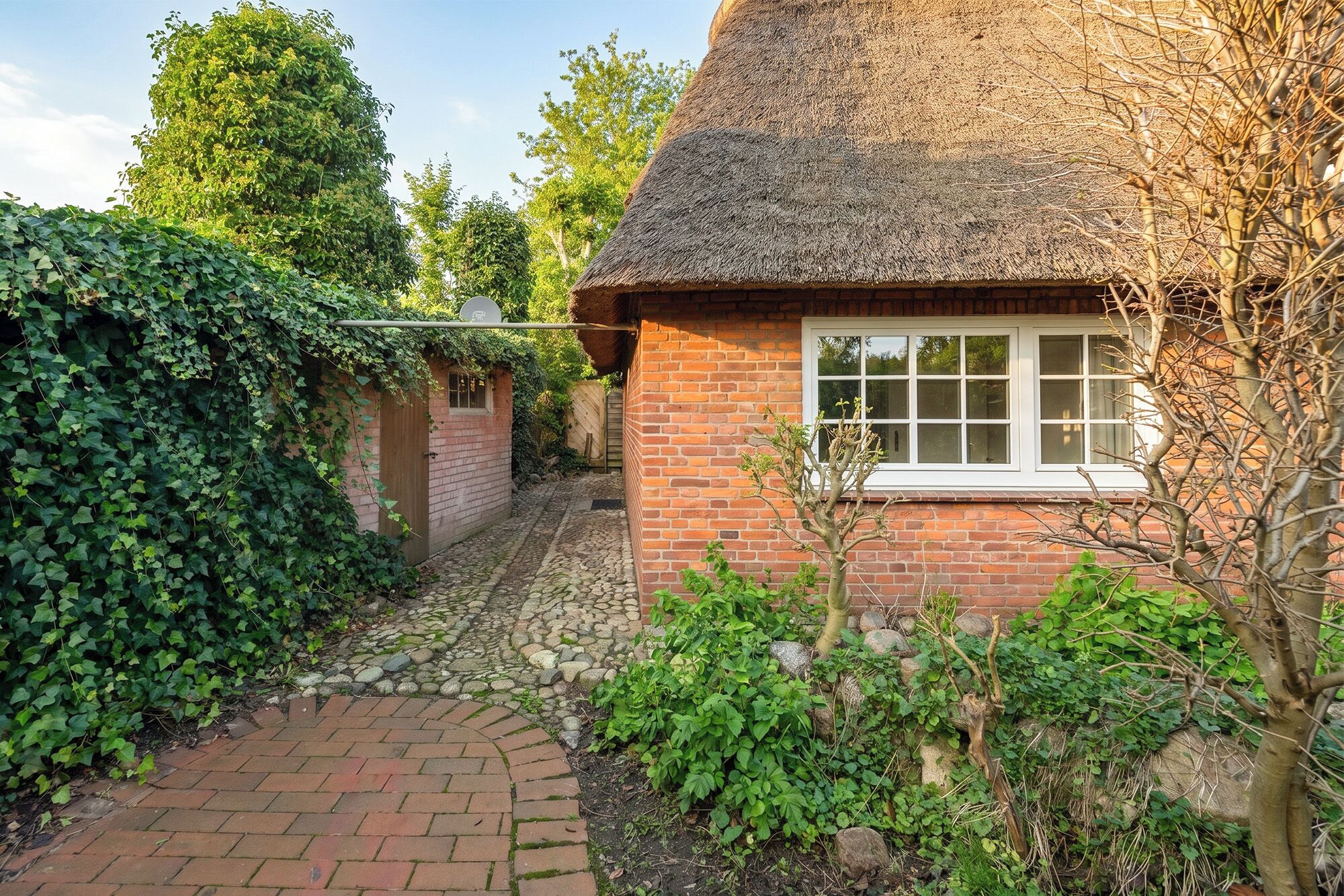 A thatched-roof cottage with a brick exterior, a narrow stone pathway, and lush greenery.