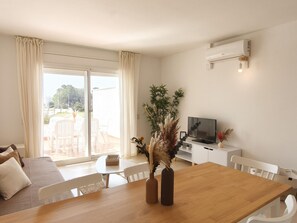 En moderne stue med et træspisebord, hvide stole, en sofa, et fjernsyn og en skydedør til en udendørs terrasse.