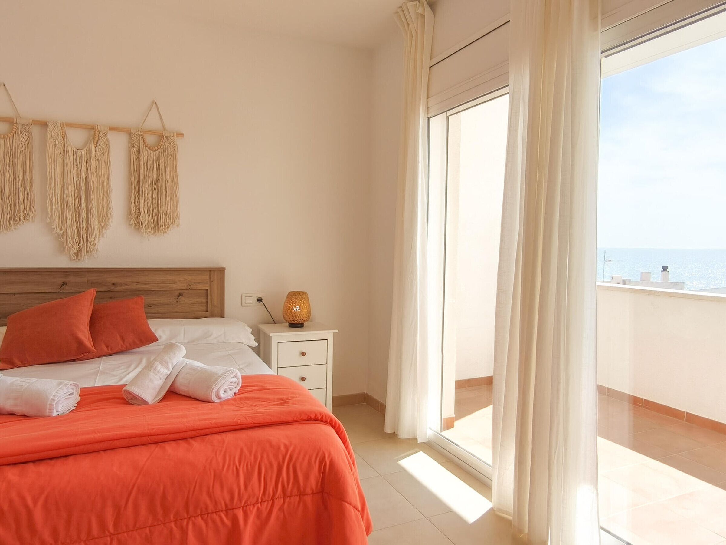 A hotel room with a bed, a nightstand, a lamp, macrame wall decor, and a view of the sea.