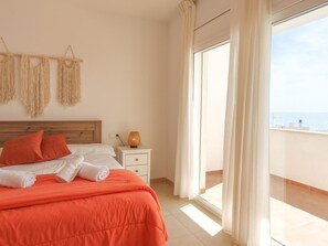 A hotel room with a bed, a nightstand, a lamp, macrame wall decor, and a view of the sea.