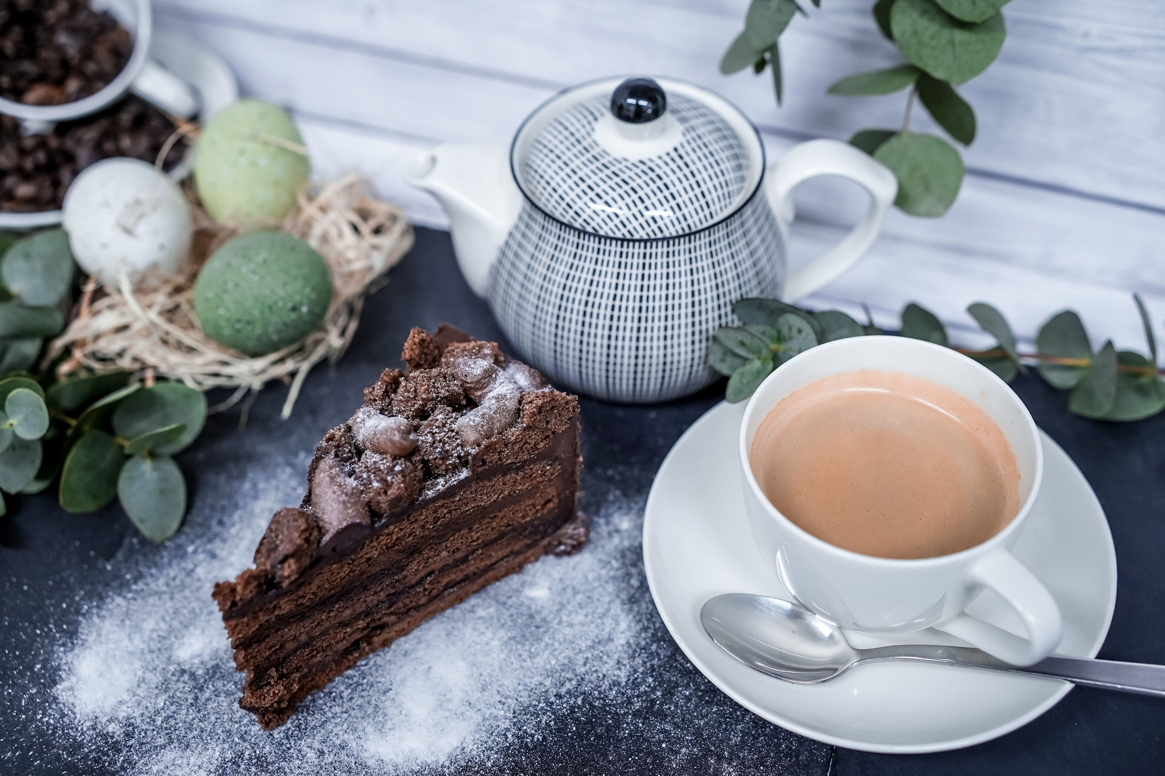 Una porción de pastel de chocolate con azúcar glas, una tetera y una taza de café sobre una mesa.