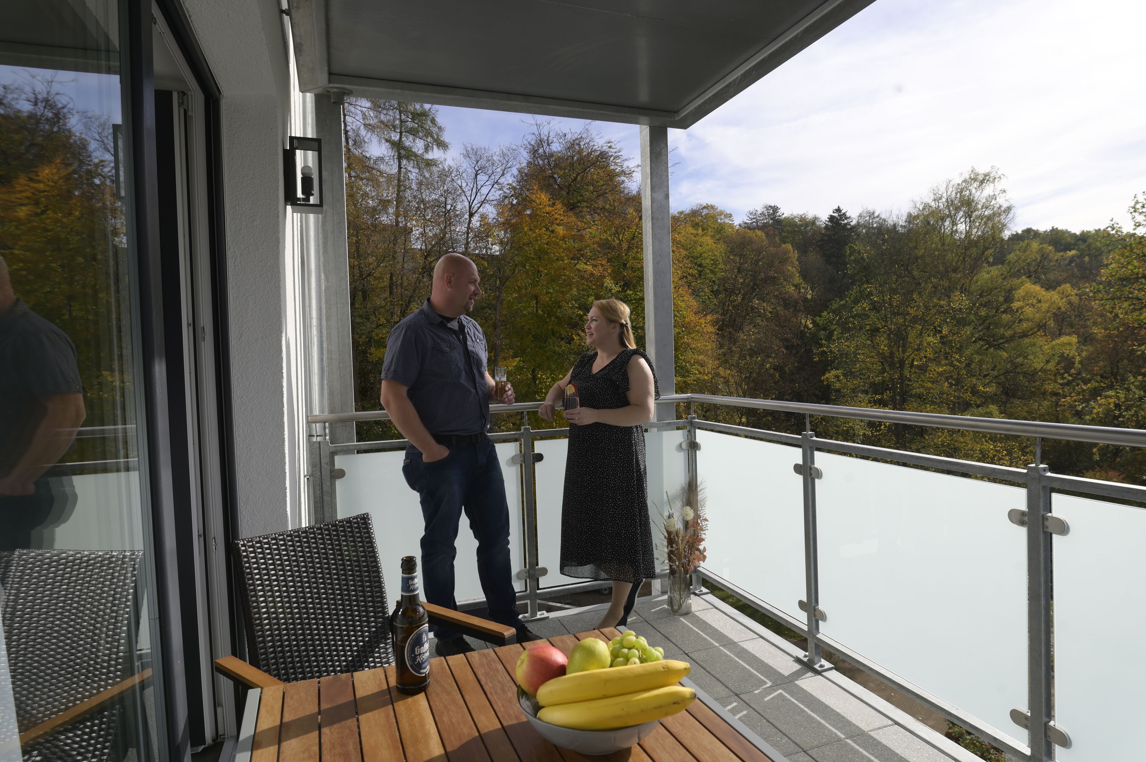 A balcony with a view of trees and a body of water. There are two people standing, a table with a bottle and a bowl of fruit, and a chair.