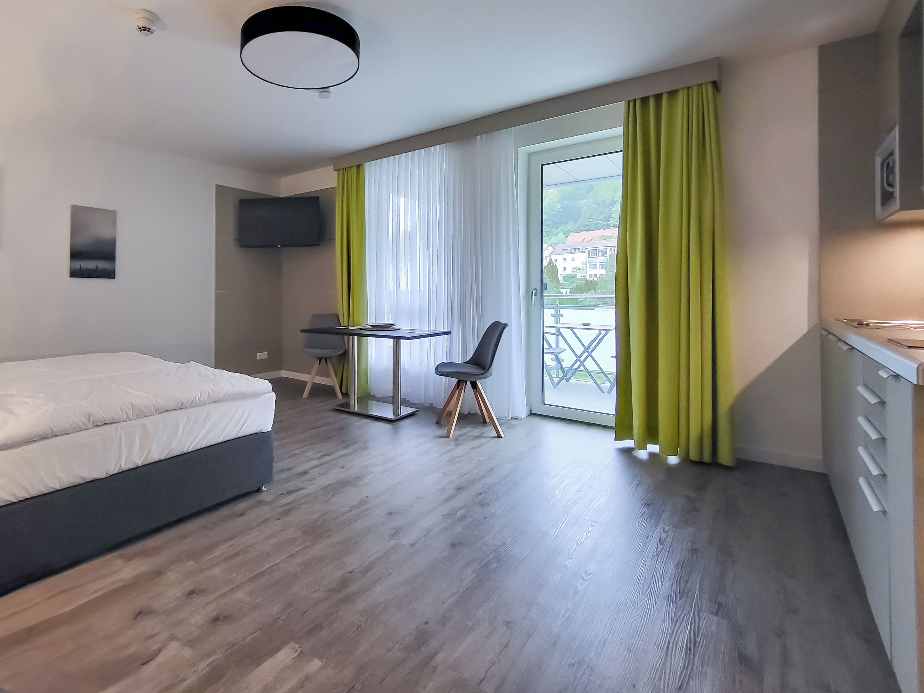 Una habitación de hotel moderna con cama, escritorio, silla, televisor y balcón con cortinas.