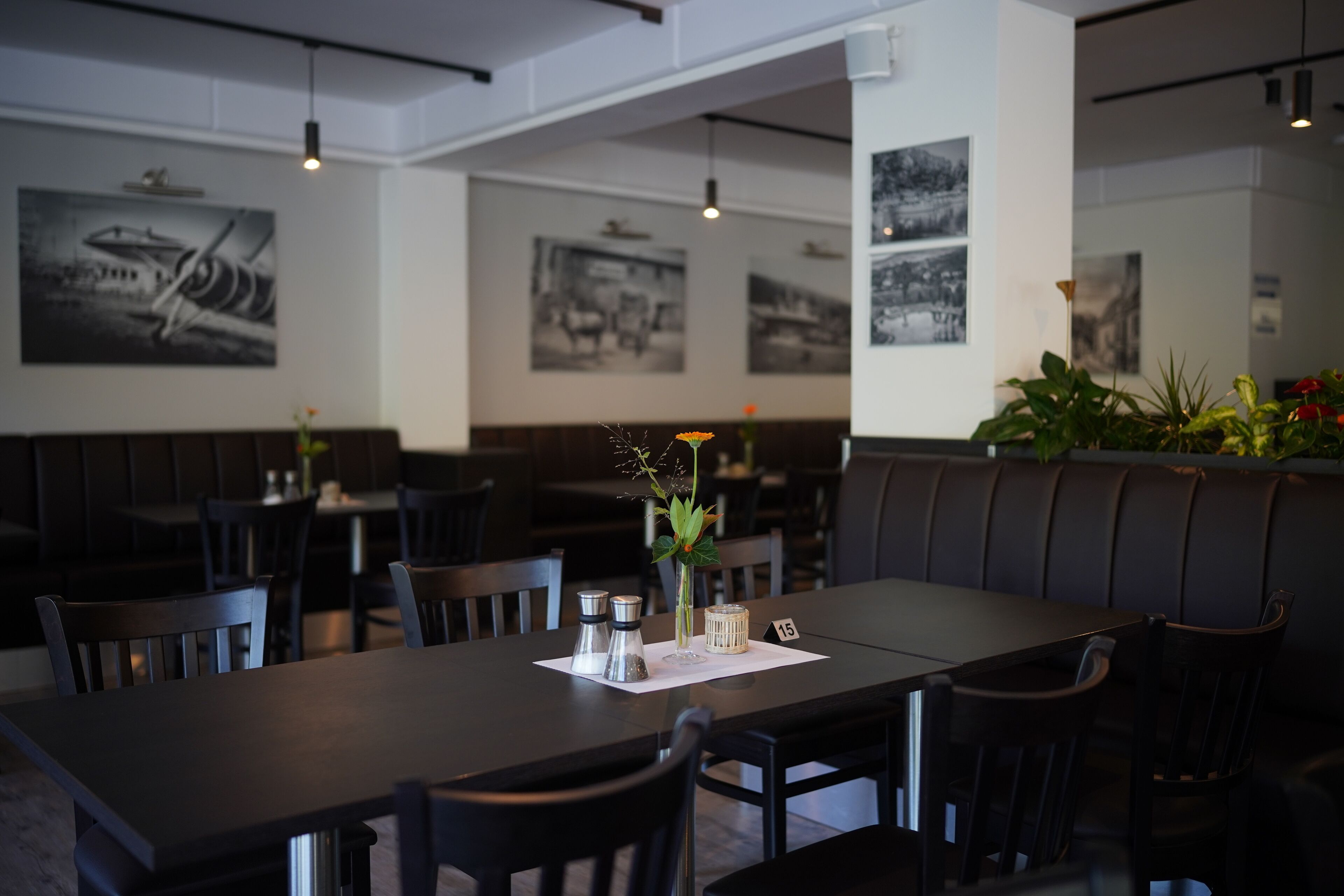 Ein Restaurant mit dunklen Holztischen und -stühlen, Schwarz-Weiß-Fotos an den Wänden und einer Zimmerpflanze auf der rechten Seite.
