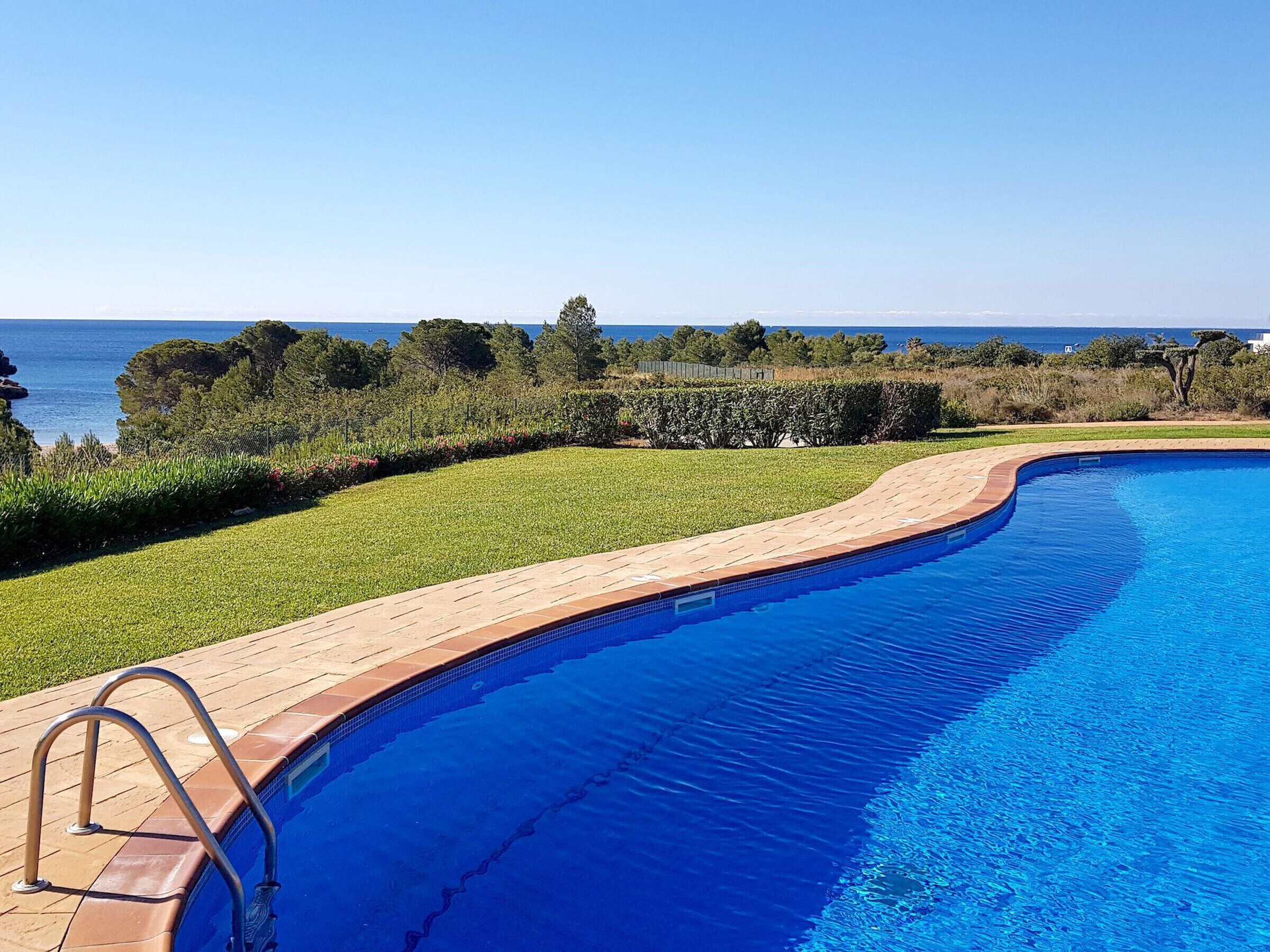 A swimming pool with a wooden deck and metal ladder, surrounded by greenery and a view of the sea.