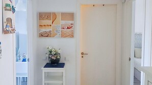 Un couloir avec une porte blanche, une petite étagère ornée d’un vase de fleurs et un collage mural composé d’images sur le thème de la plage.