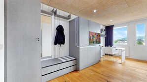 A modern, well-lit room with a wooden floor, a desk, a chair, and a large window offering a view of buildings and greenery. - Evi's Home Hotel & Apartments (Bern)