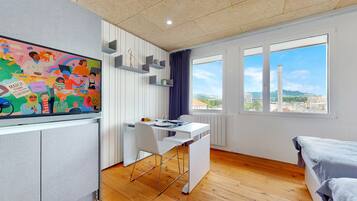 A modern room with a desk, a chair, a bed, a large window with a city view, and a colorful mural on the wall.