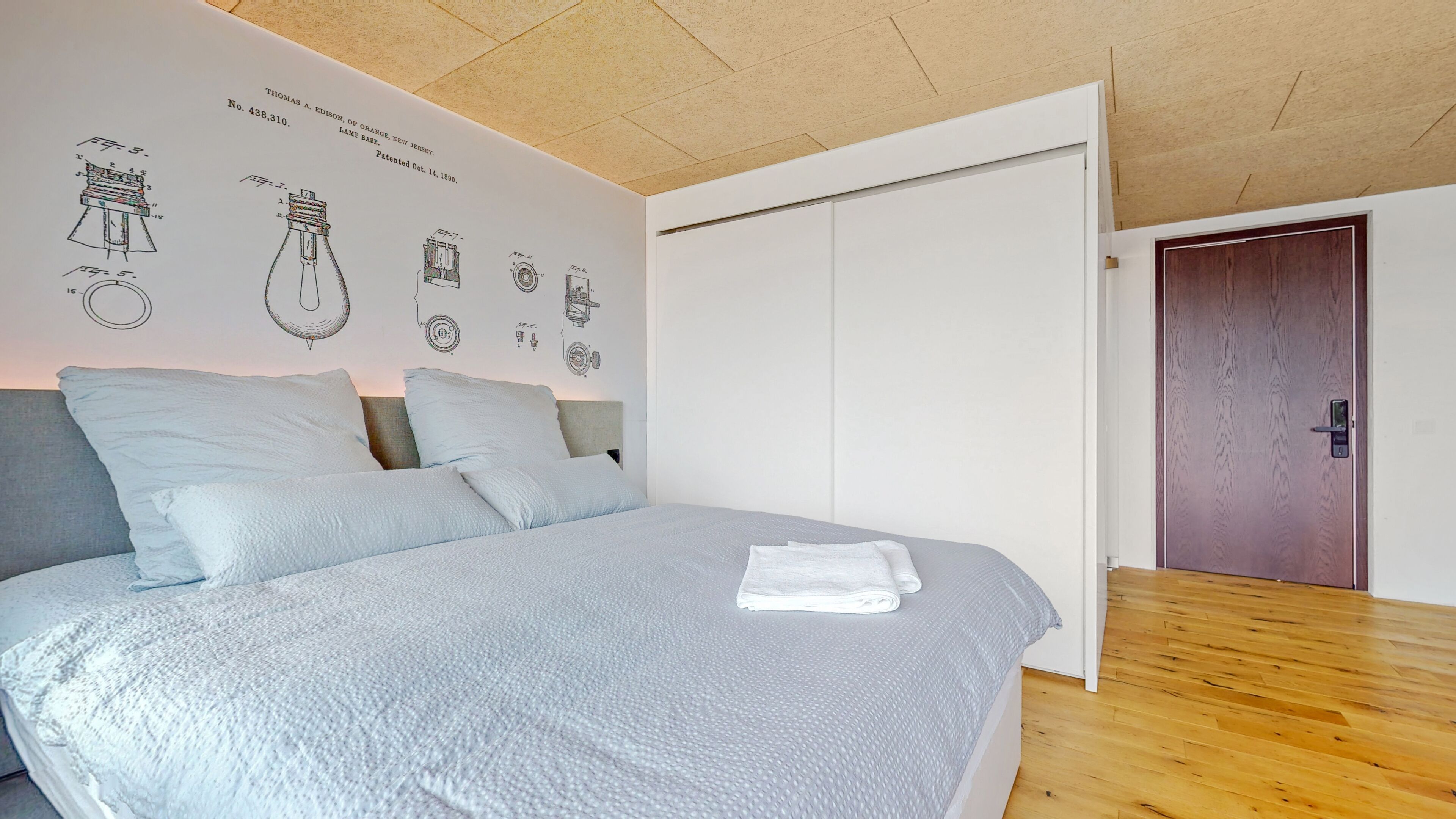 Un dormitorio con cama, almohadas, una pared con dibujos técnicos, una puerta de madera y un techo con vigas de madera.
